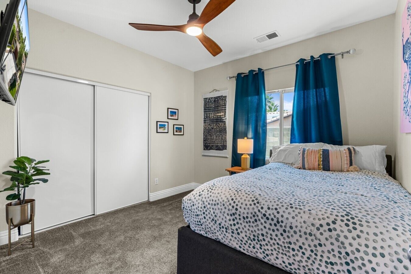 A bedroom with a bed, a ceiling fan, a nightstand, a lamp, a window with curtains, and a wall with framed pictures.
