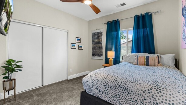 A bedroom with a bed, a ceiling fan, a nightstand, a lamp, a window with curtains, and a wall with framed pictures. - Entire Home + Private Casita | Private Pool & Spa | Putting Green | Fully Fenced Yard| Community Amenities (Palm Springs)
