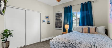 Un dormitorio con cama, ventilador de techo, mesita de noche, lámpara, ventana con cortinas y un cuadro en la pared.