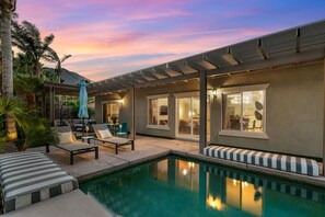 Ein moderner Poolbereich im Freien mit Liegestühlen, einer überdachten Lounge und Blick auf das Haus.