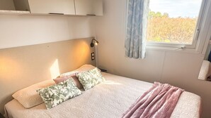 A bedroom with a bed, pillows, a blanket, a bedside lamp, a window with curtains, and a headboard. - Homerez - Bungalow in Saint-Raphaël (Saint-Raphaël)