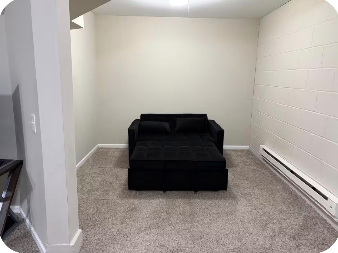Una habitación pequeña con alfombra y un sofá cama negro frente a una pared lisa.