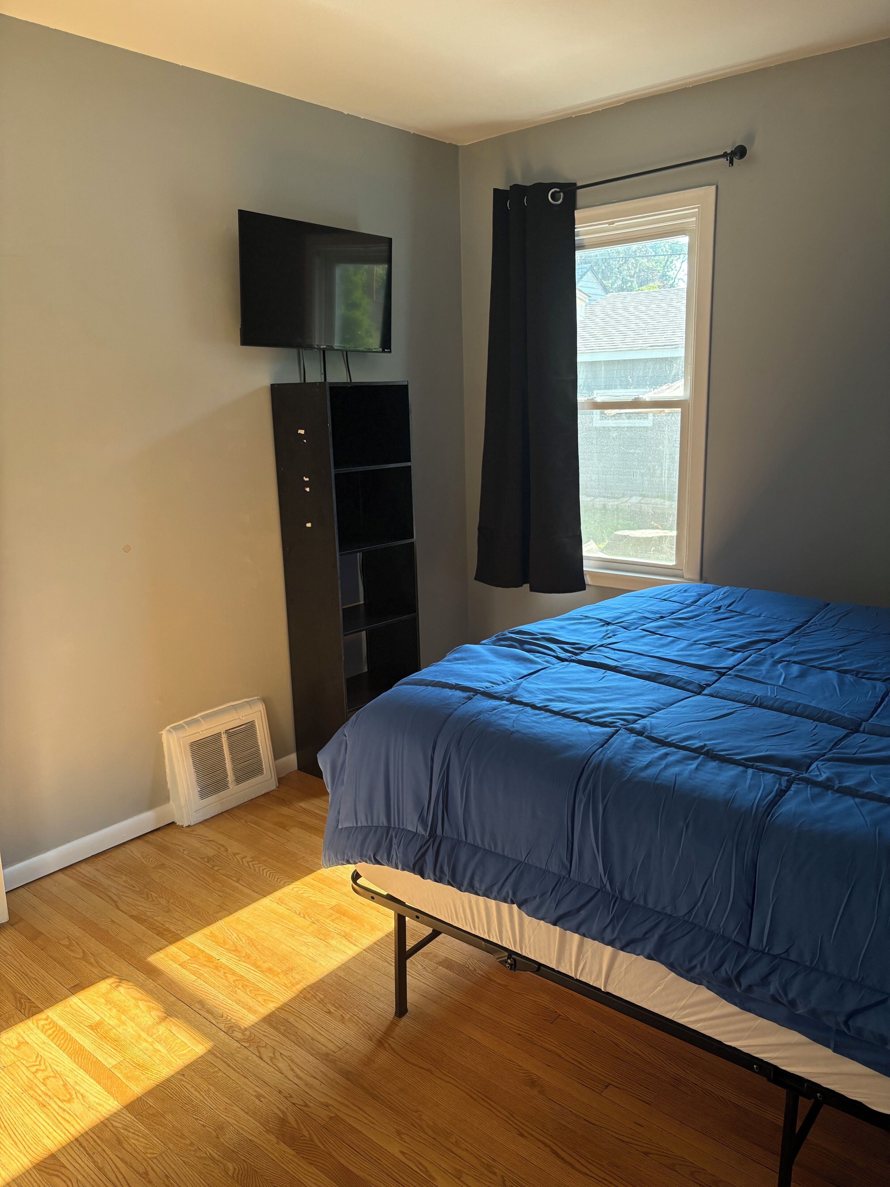 A bedroom with a bed, a TV mounted on the wall, a window with a curtain, and a radiator.