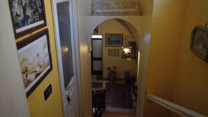 Um corredor estreito com uma porta à esquerda, um arco decorativo no centro e uma pequena escadaria que leva a um andar superior.