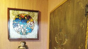 Quarto com porta de madeira com padrões decorativos, uma janela de vitrais acima de uma estátua, e uma pequena mesa com um lampião verde.