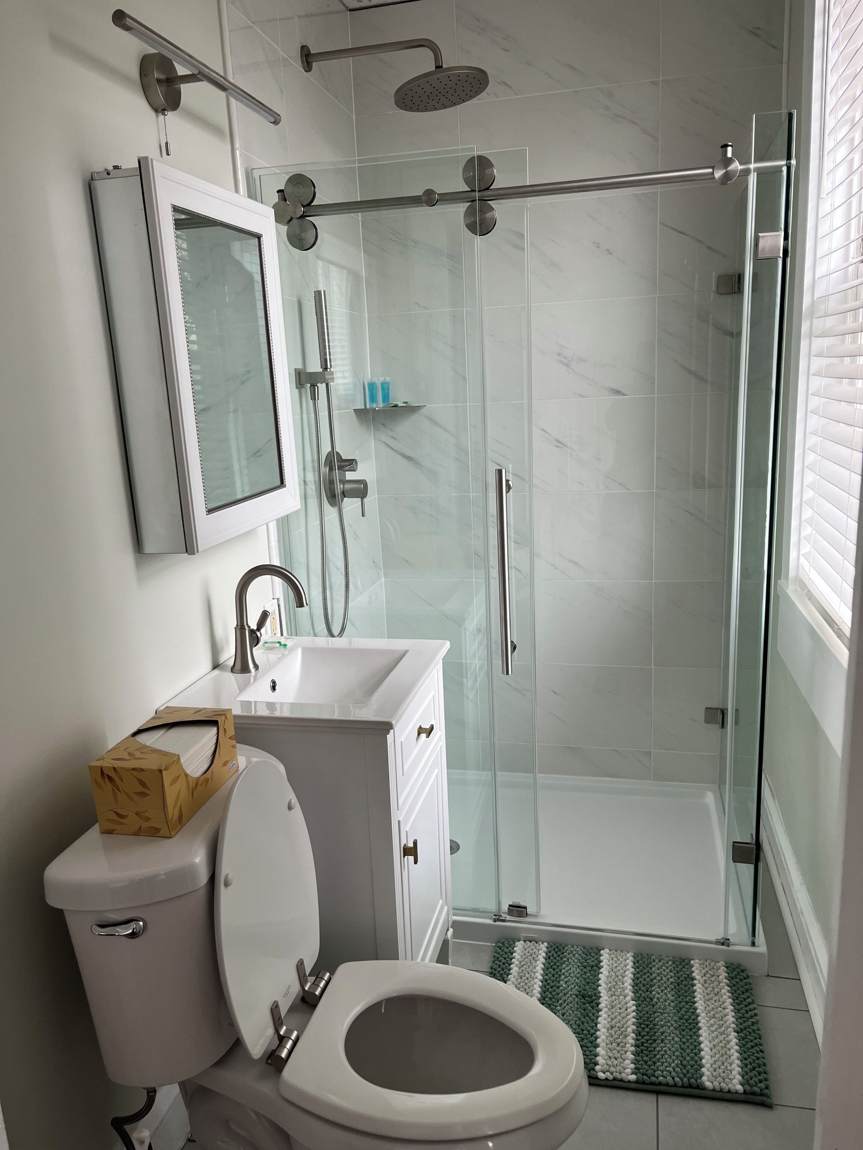 Une salle de bain moderne dotée d’une douche à parois de verre, de toilettes, d’un lavabo avec un miroir et d’une fenêtre laissant entrer la lumière naturelle.