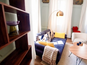 Ein modernes Wohnzimmer mit einem dunkelblauen Sofa, gelben Kissen, einer gestreiften Decke, einer Stehlampe und einem kleinen runden Tisch mit zwei blauen Tassen.