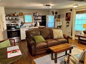 A compact living space with a kitchenette, a brown leather sofa, a wooden coffee table, and a dining area with a chair. - Two Cottages in the Woods! 4 mins to Dahlonega Square (Dahlonega)