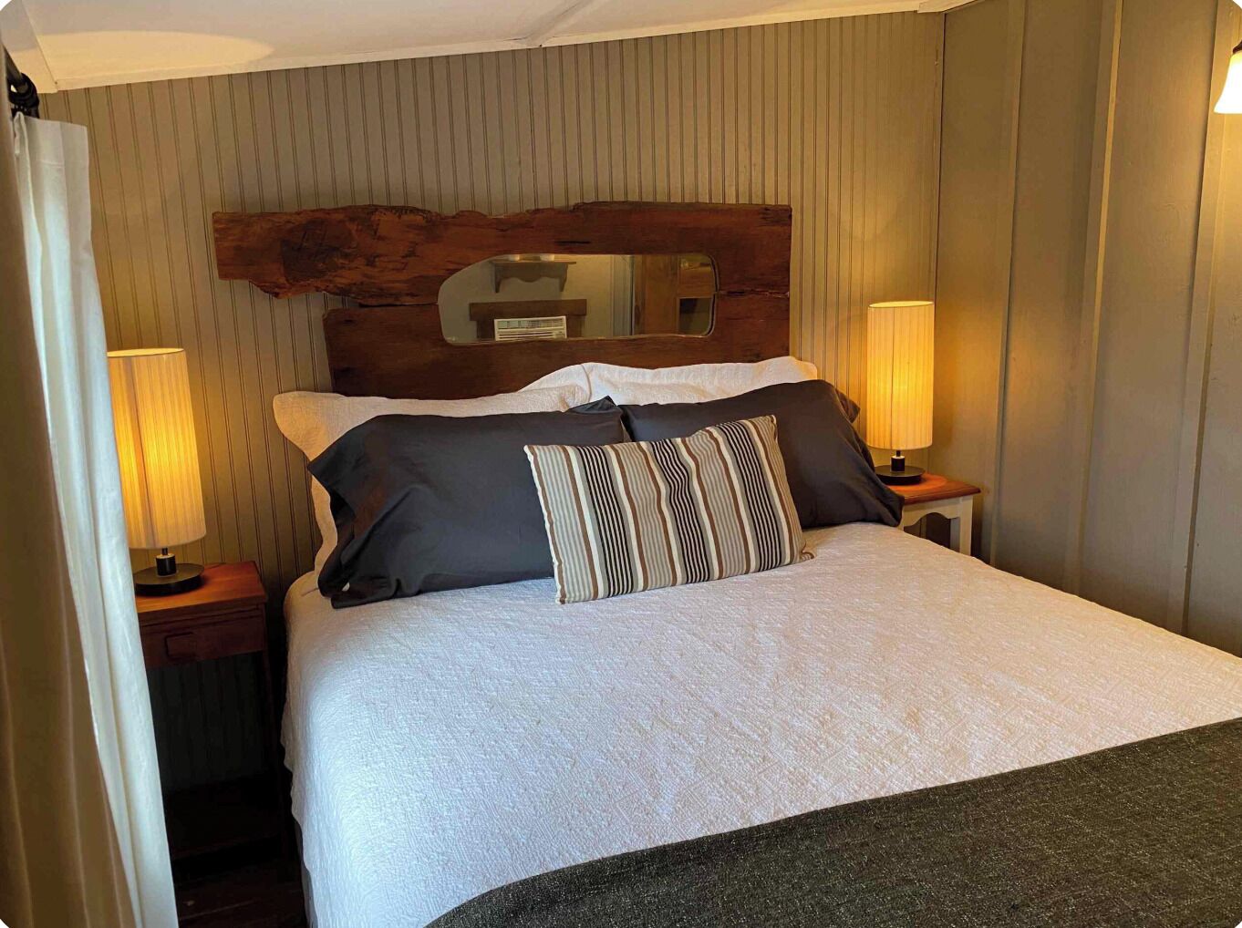 Une chambre d’hôtel avec un lit, deux lampes de chevet, une tête de lit agrémentée d’un miroir et un rideau à moitié tiré.