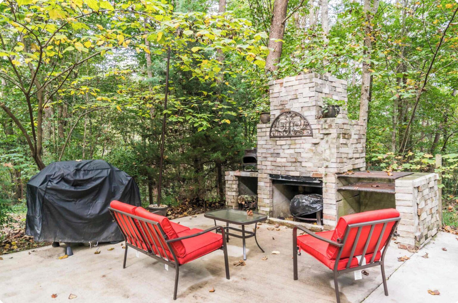 Une terrasse extérieure dotée d’une cheminée en pierre, de deux fauteuils rouges avec coussins et d’un barbecue couvert.