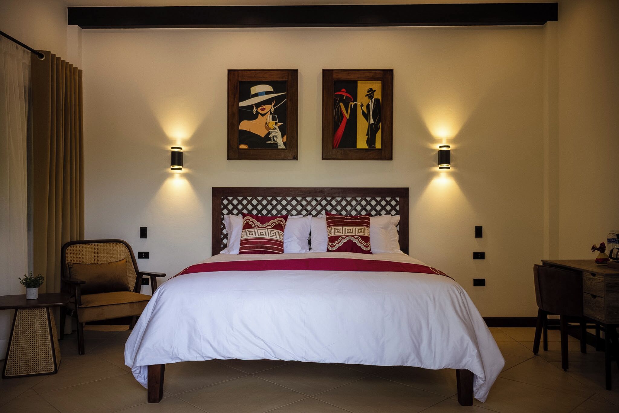 A hotel room with a large bed, two wall-mounted lamps, a chair, a small table, and two framed artworks above the bed.