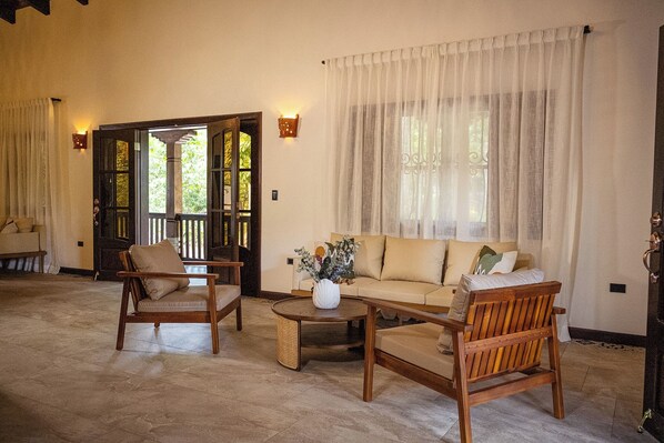 A room with a sofa, two wooden chairs, a coffee table, and a door leading outside.