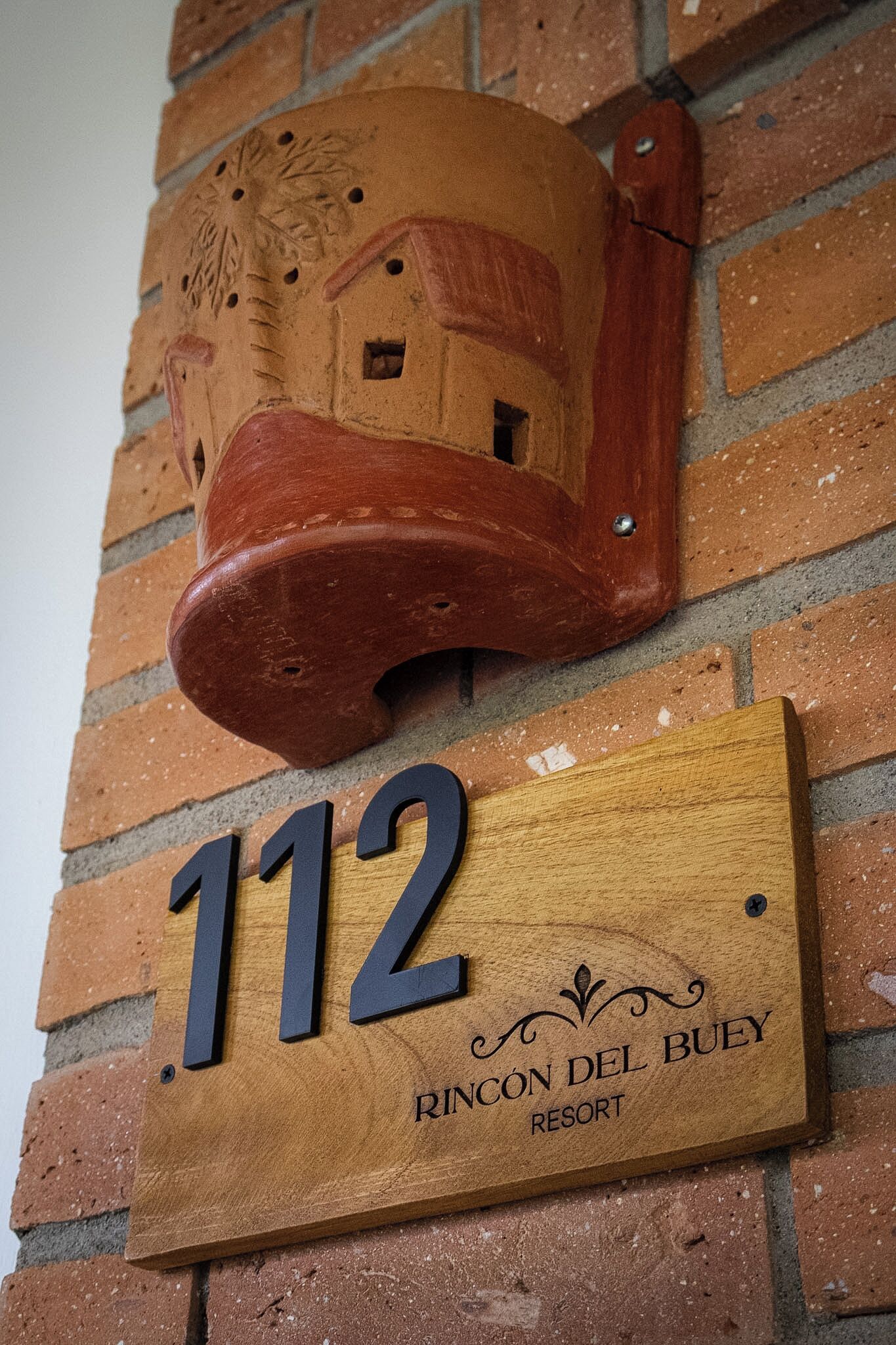 Placa de madeira com o número 112 e o texto 'Rincon Del Buey Resort' em uma parede de tijolos.