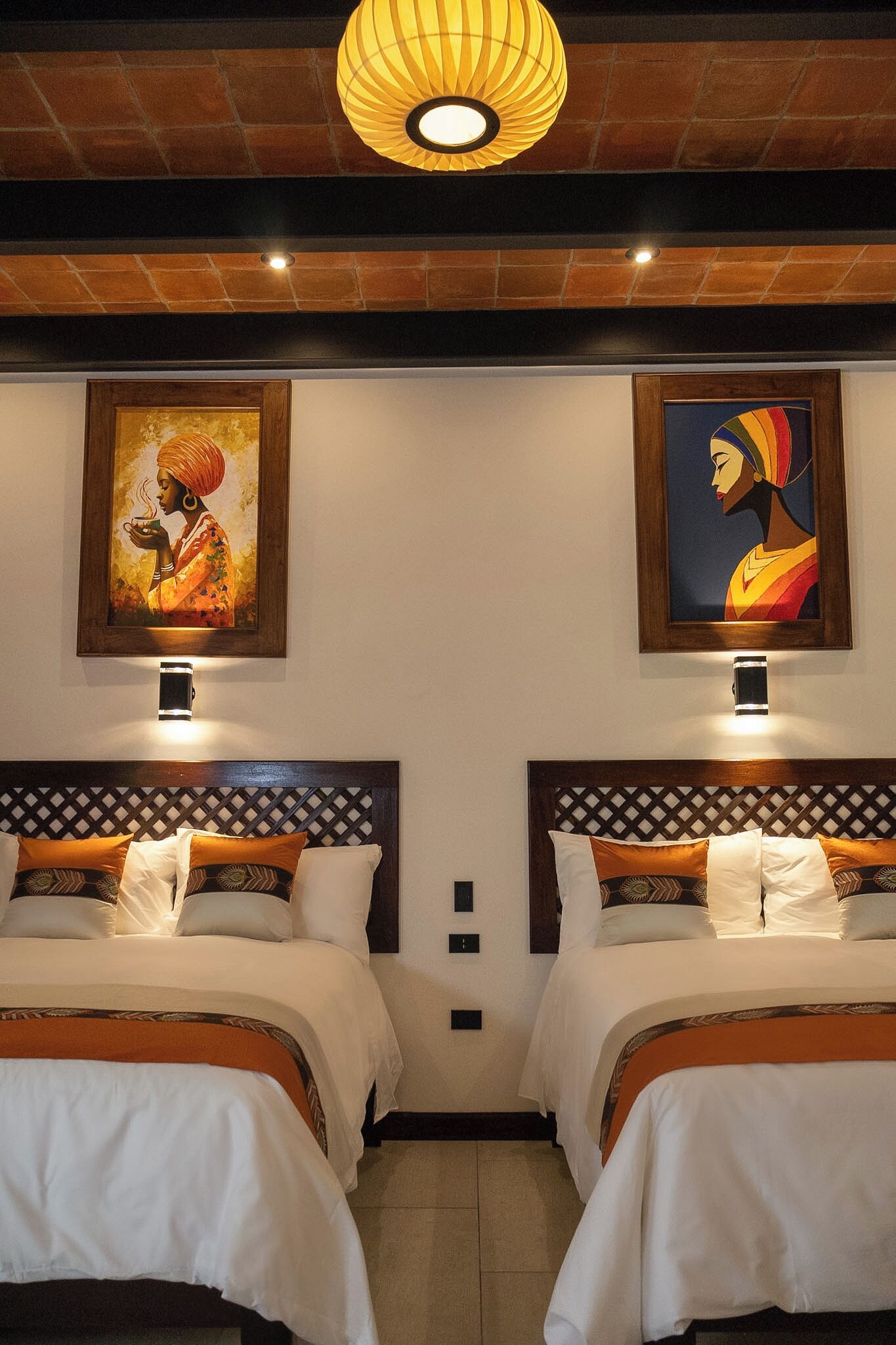 Ein Hotelzimmer mit zwei Betten, jedes mit einem Kopfende mit Rautenmuster. Über jedem Bett befinden sich wandmontierte Leuchten, und zwischen den Betten hängt ein Kunstwerk in einem Bilderrahmen.