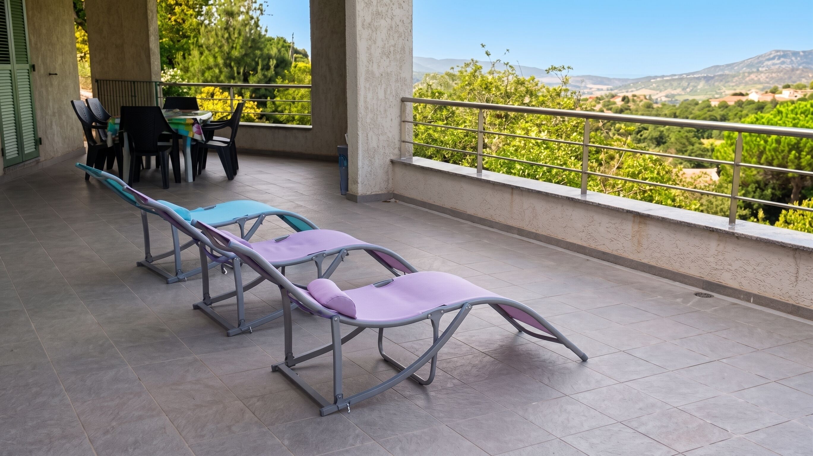 Un balcon agrémenté de chaises longues, d’une table et de chaises, offrant une vue sur un paysage verdoyant avec des collines à l’horizon.