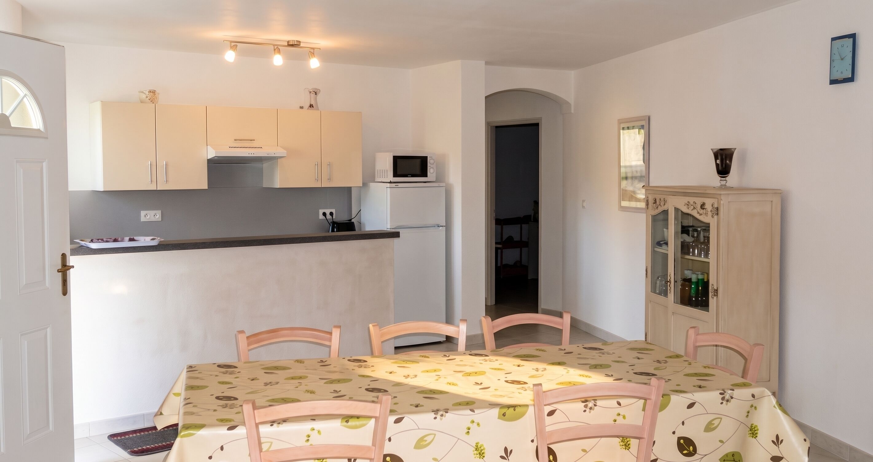 Un coin repas avec une table dressée pour quatre personnes, une cuisine équipée de placards et d’appareils électroménagers, et un meuble avec des portes en verre.