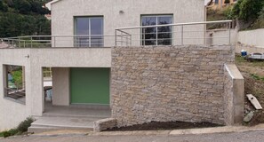 A modern building with a stone facade, a green garage door, and a metal balcony railing. - Homerez - House in Oletta, sea/mountain view. (Oletta)