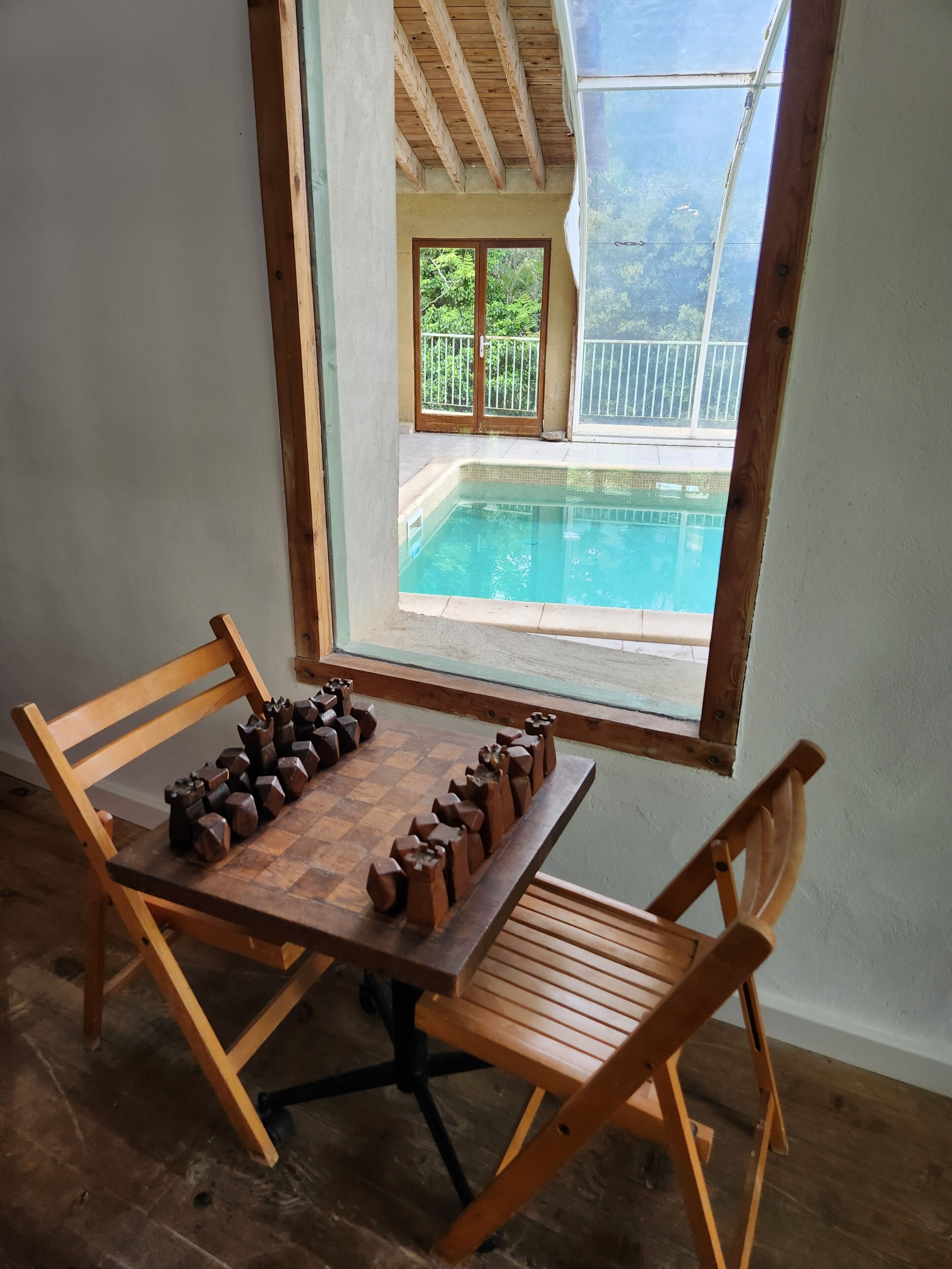 Une table en bois avec des pièces d’échecs, une fenêtre donnant sur une piscine et un plancher en bois.