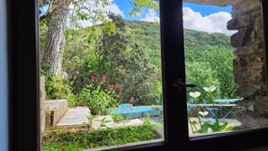 A window view of a lush garden with a stone wall, a tree, and outdoor furniture. - Secluded vacation rental with a pool in the heart of the Corbières – Nature & Relaxation (Lairière)