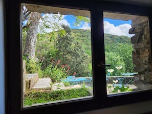 A window view of a lush garden with a stone wall, a tree, and outdoor furniture. - Secluded vacation rental with a pool in the heart of the Corbières – Nature & Relaxation (Lairière)