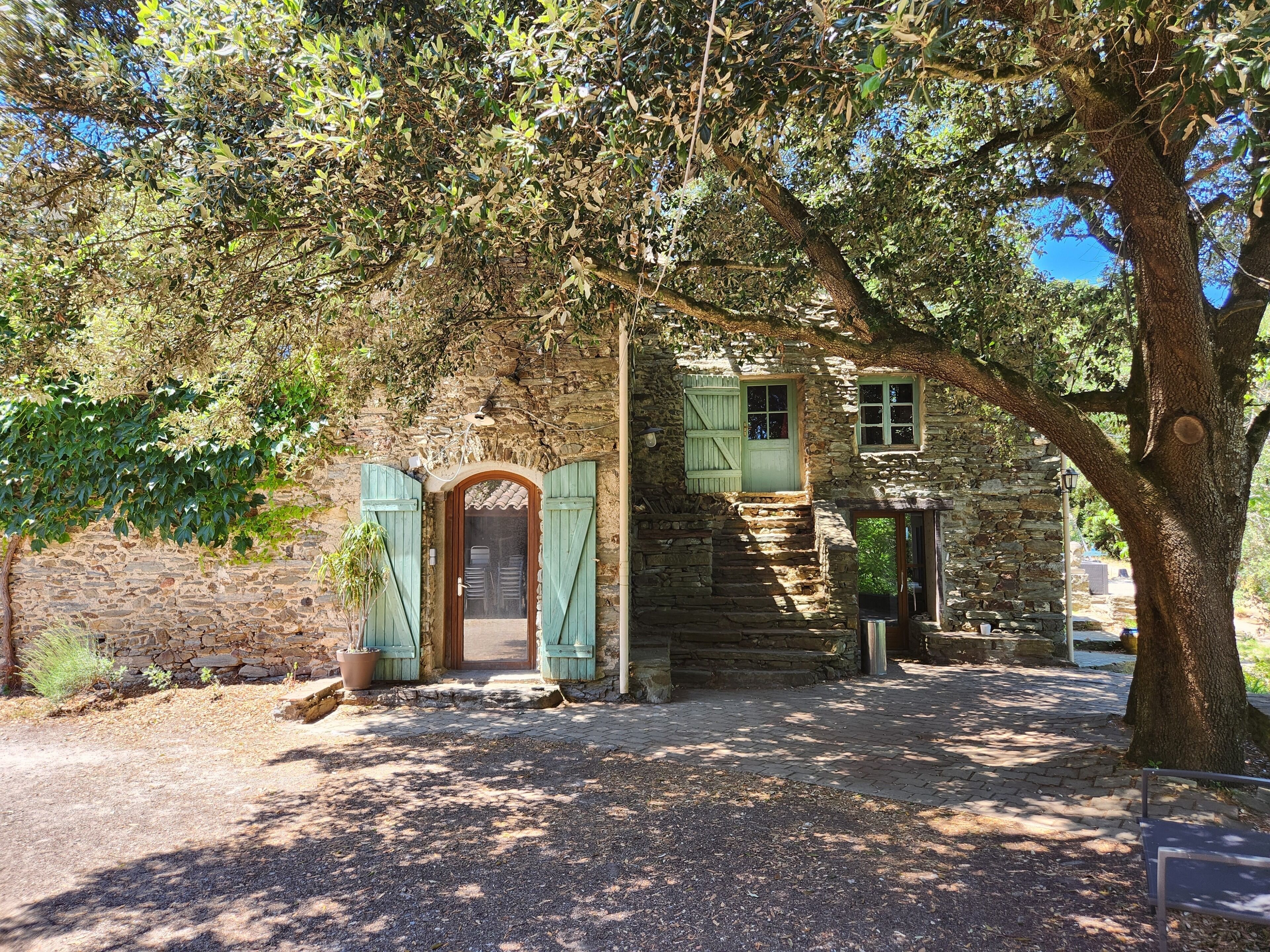Un bâtiment rustique en pierre, doté d’une porte et de volets en bois, entouré d’arbres et bordé d’un chemin de gravier.