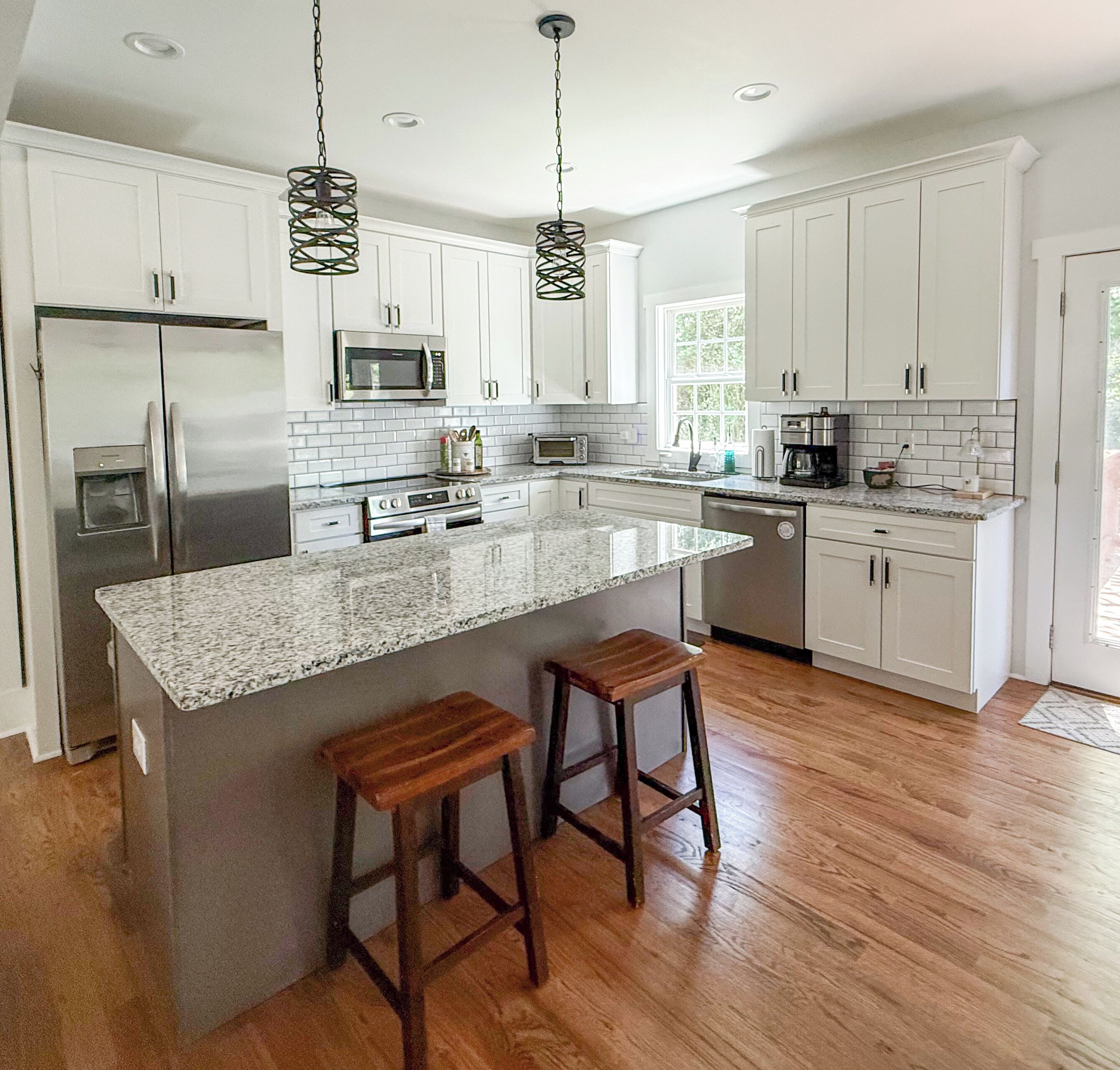 Una cocina moderna con muebles blancos, electrodomésticos de acero inoxidable, encimera de granito y dos taburetes de madera.