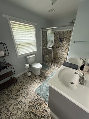 A bathroom with a stone-patterned floor, a white sink, a toilet, and a shower area. - Cozy Creekside Cottage – AC, Dock, Kayaks & Firepit (Deltaville,)