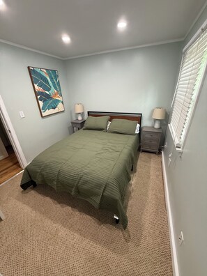 A bedroom with a bed, two nightstands, a wall-mounted lamp, a painting, and a window with blinds. - Cozy Creekside Cottage – AC, Dock, Kayaks & Firepit (Deltaville,)