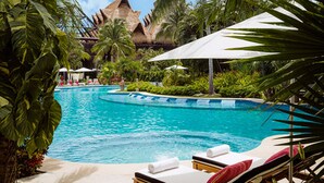 Ein Swimmingpool mit Liegestühlen und Sonnenschirmen, umgeben von tropischer Vegetation und einer Struktur mit einem Reetdach.