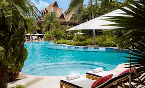 Ein Swimmingpool mit Liegestühlen und Sonnenschirmen, umgeben von tropischer Vegetation und einer Struktur mit einem Reetdach.