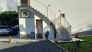 Una casa blanca de dos pisos con una escalera que lleva al piso superior, un pequeño balcón y un jardín con plantas en macetas.