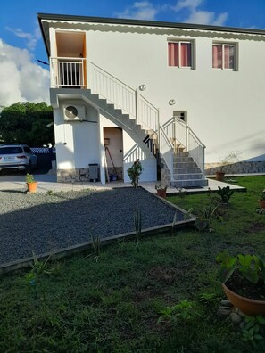 A white two-story house with a staircase leading to the upper floor, a small balcony, and a garden area with potted plants.