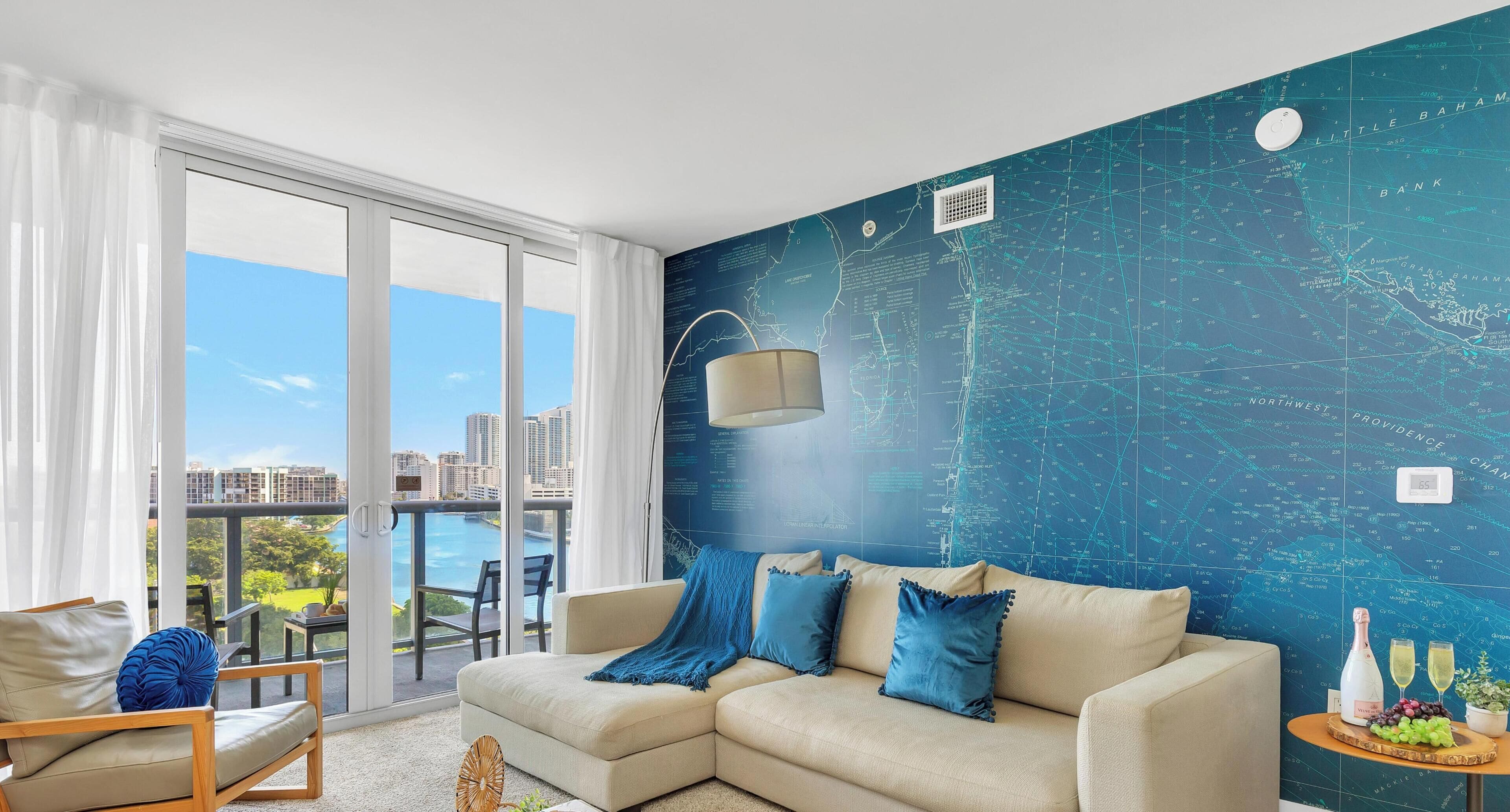 A modern living room with a large window, a beige sofa, and a blue wall with a map design.