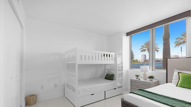 A bedroom with a bunk bed, a bedside table with a lamp, a window with a view of palm trees and buildings, and a green throw on the bed.