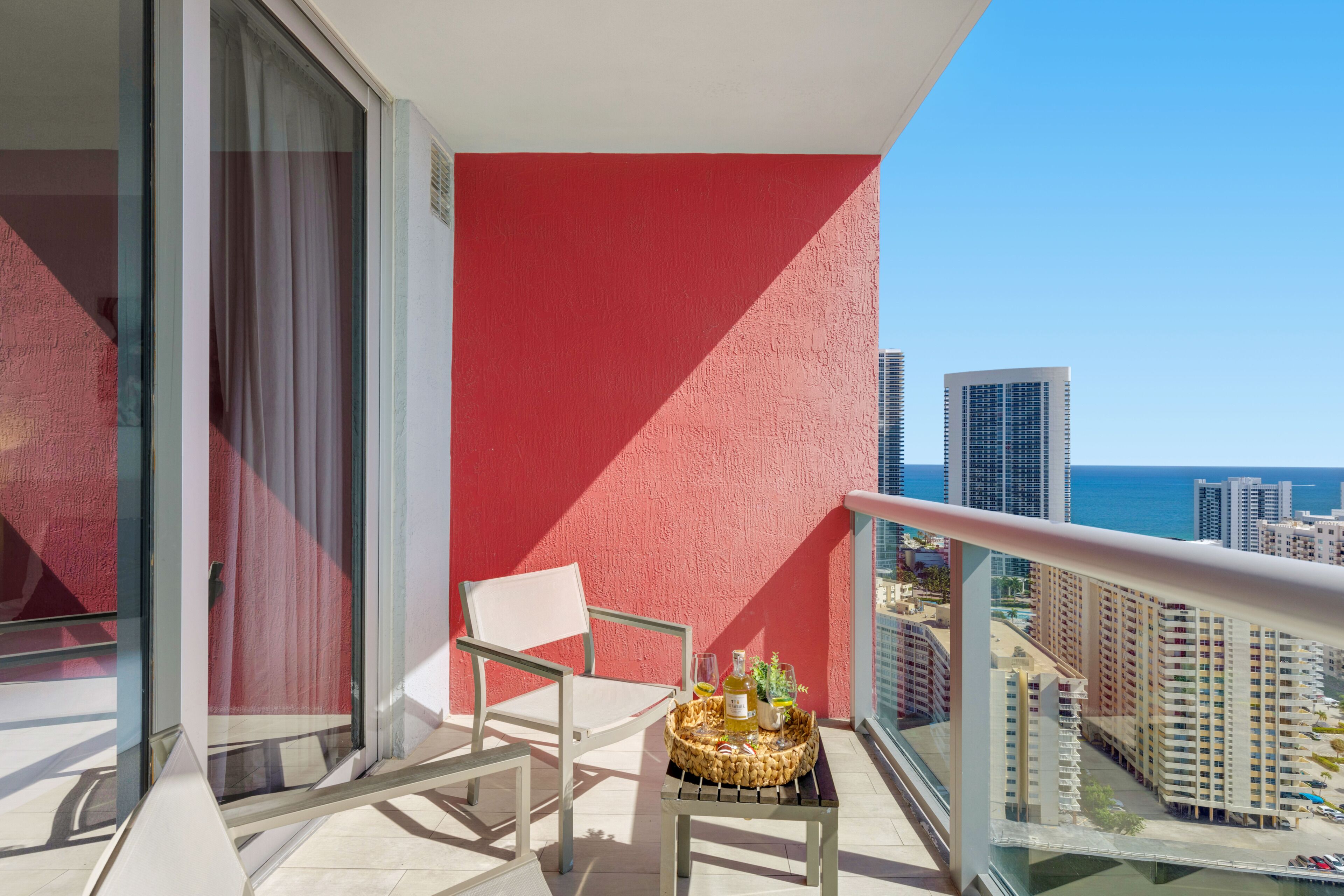 A balcony with a chair, a small table with a basket, and a view of the ocean and buildings.