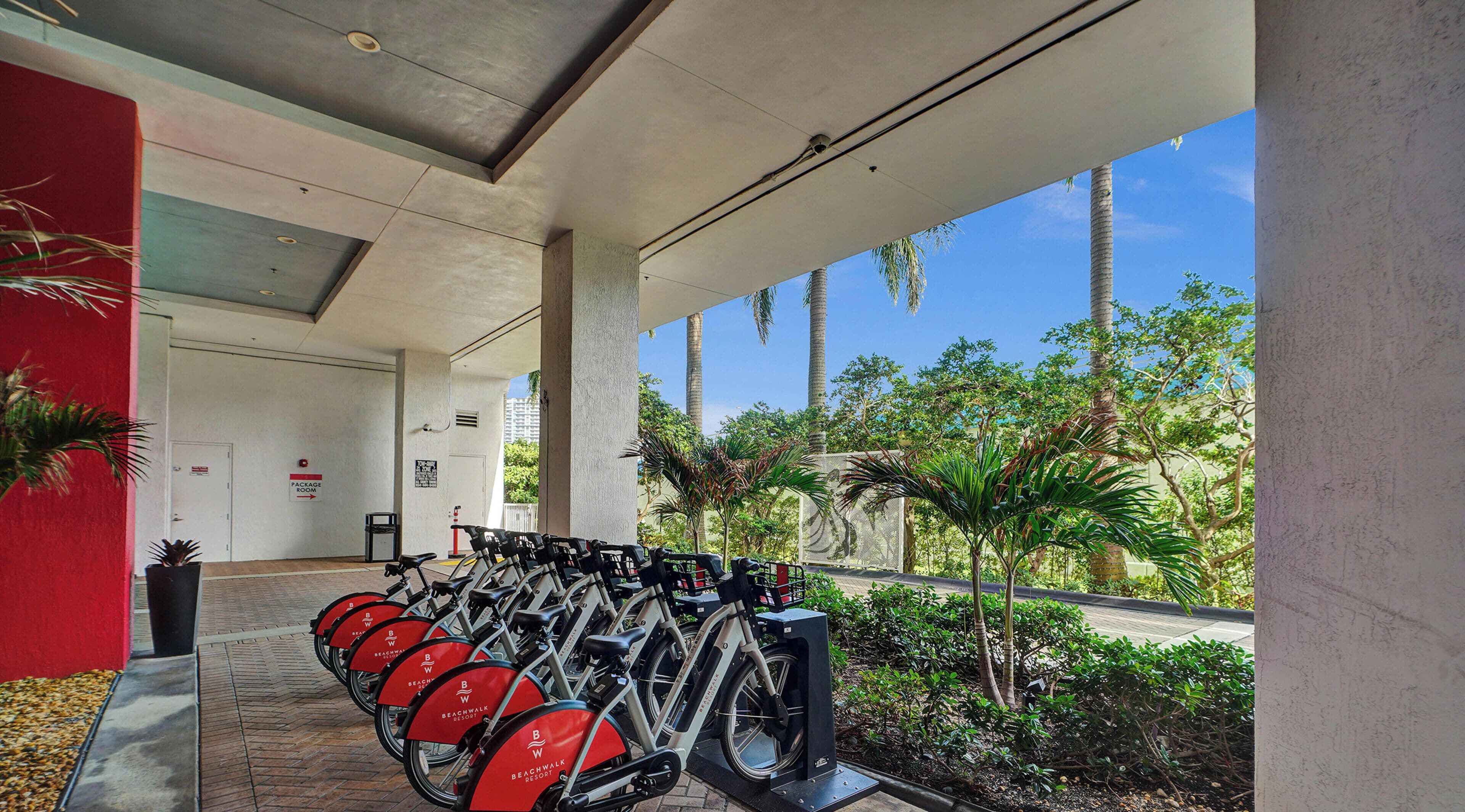 A covered bike rental area with multiple bicycles, a pathway leading to a building, and a view of palm trees and greenery.