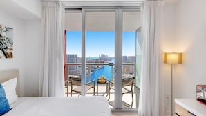 A hotel room with a bed, a desk, a chair, a table with a basket, and a view of a cityscape and water.