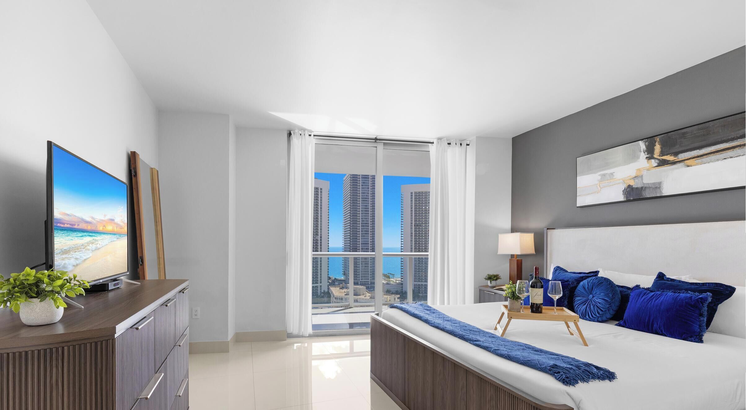 A modern bedroom with a bed, a TV, a desk, and a view of high-rise buildings.
