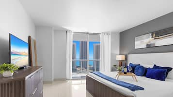A modern bedroom with a bed, a TV, a desk, and a view of high-rise buildings.