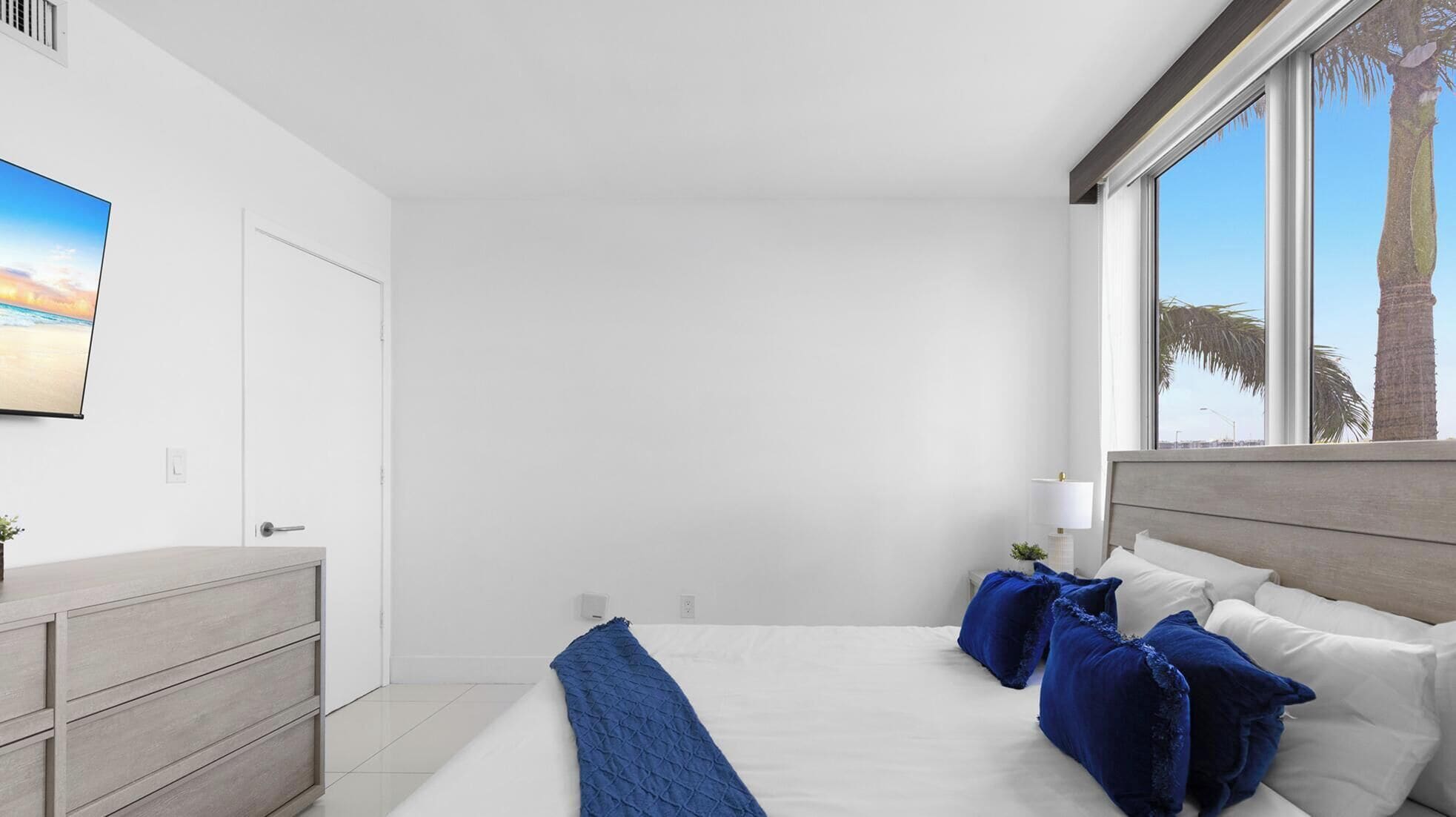 A bedroom with a bed, a nightstand, a lamp, and a window with a view of palm trees.