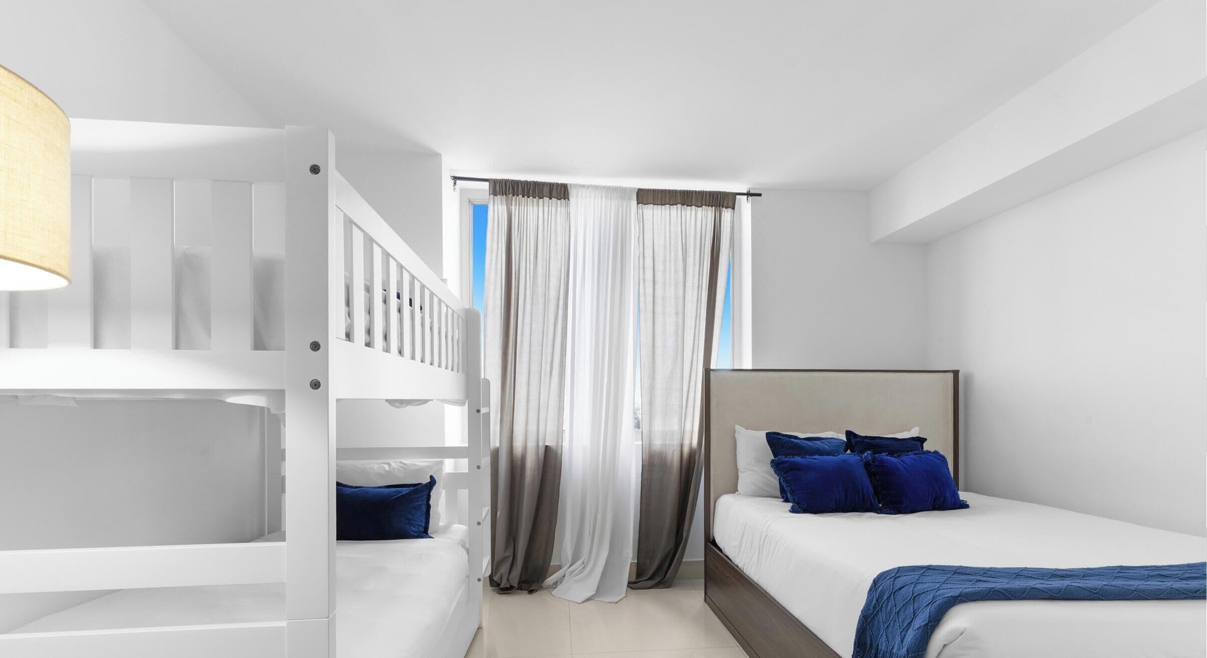 A bedroom with a bunk bed, a single bed with blue pillows, and a window with sheer curtains.