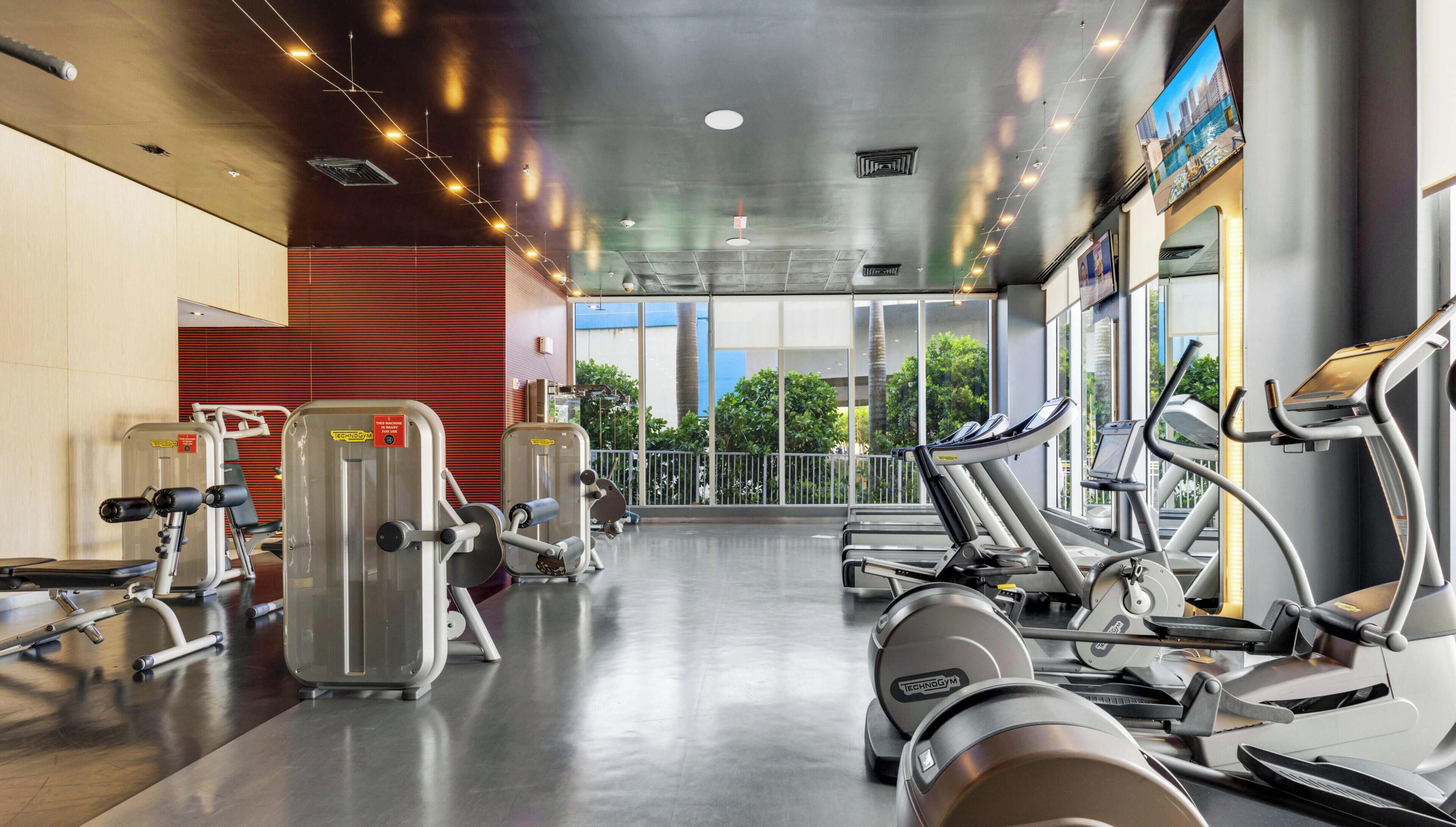 Une salle de sport équipée de divers appareils d’exercice, dotée de grandes fenêtres et offrant une vue sur la verdure environnante.