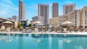A poolside area with lounge chairs and umbrellas, a clear blue pool, and high-rise buildings in the background.