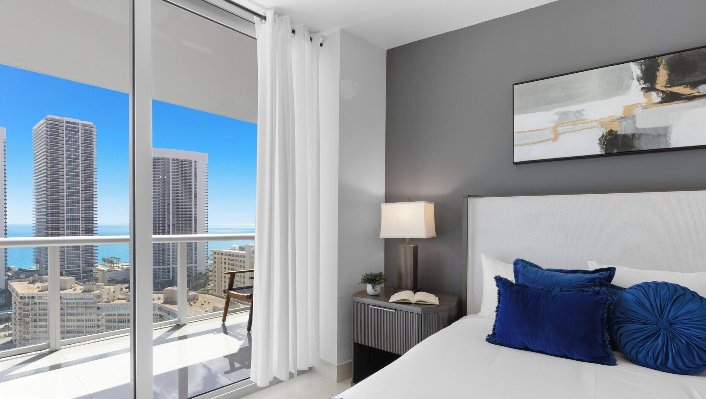 A modern hotel room with a large window, a bed with blue pillows, a bedside table with a lamp, and a painting on the wall.