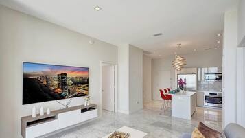A modern living room with a large flat-screen TV, a kitchen area, and a dining space.
