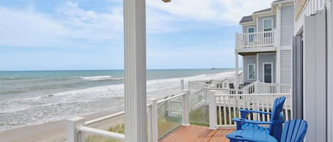 A wooden deck with a view of the ocean, a balcony with blue chairs, and a building with a white railing.