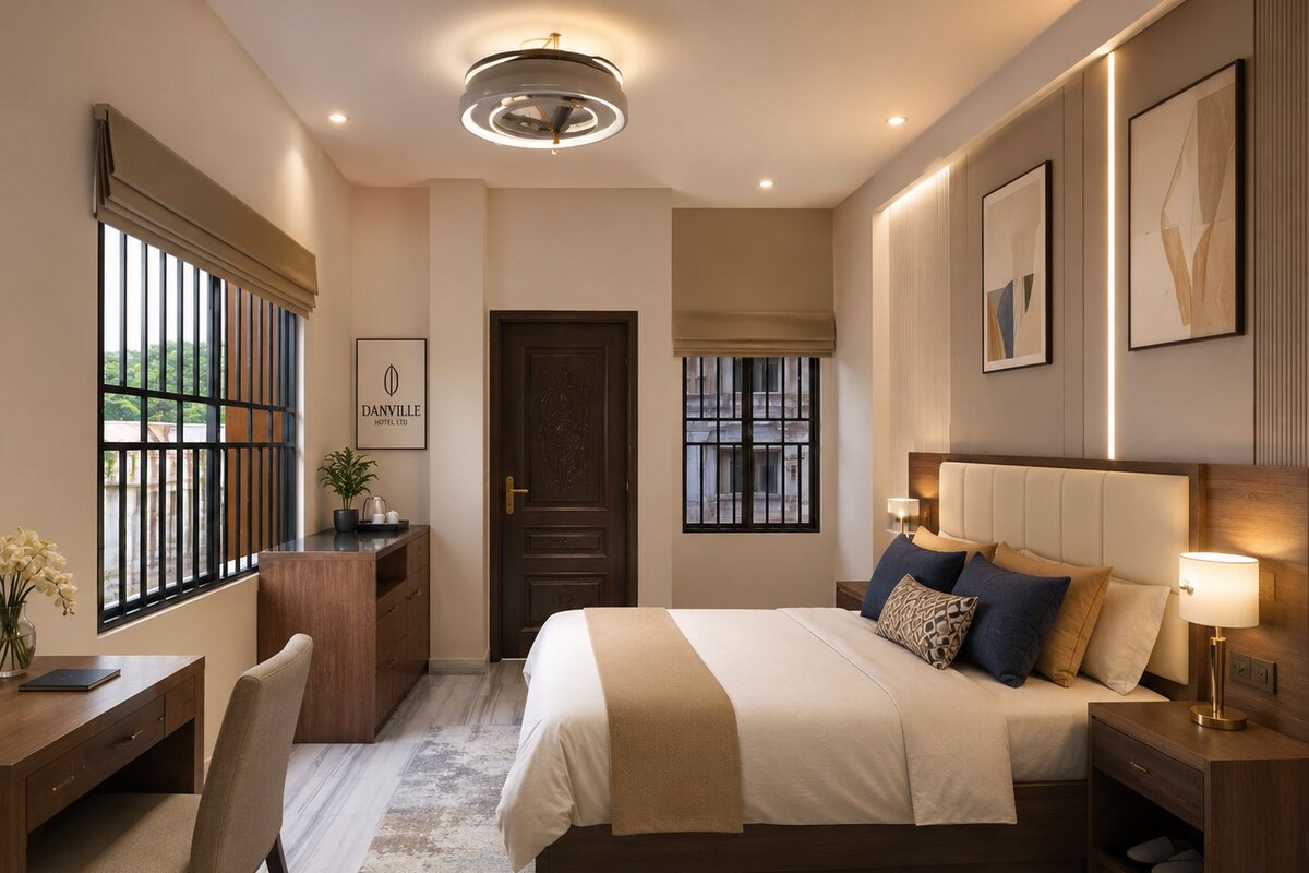 Quarto de hotel com cama, mesa de cabeceira, abajur, escrivaninha, cadeira e vista para o exterior pela janela.