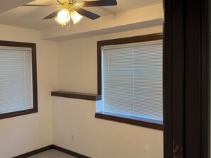 Ein Zimmer mit zwei Fenstern, einem Deckenventilator und einem an der Wand befestigten Regal.