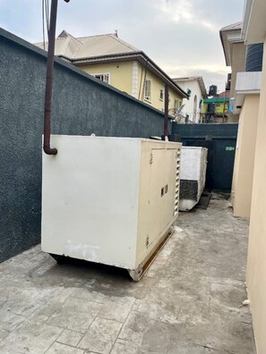 A narrow alleyway with a large white industrial unit and a black gate, surrounded by buildings. - RT24 Hotel (Lagos)