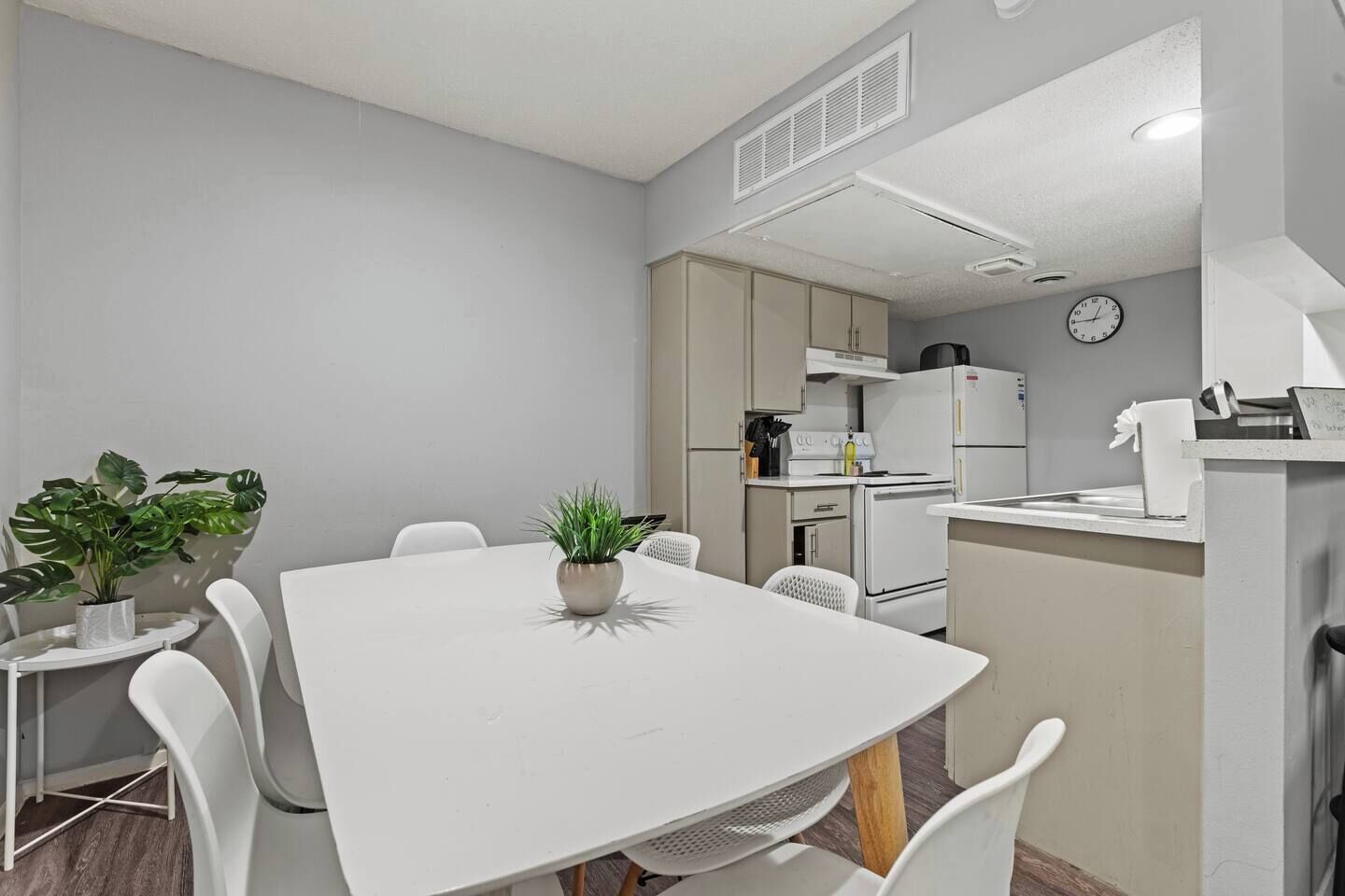 Une cuisine compacte avec un réfrigérateur, une cuisinière et un four à micro-ondes, tous de couleur blanche. On y trouve un petit coin repas avec une table et des chaises blanches, ainsi que deux plantes en pot.